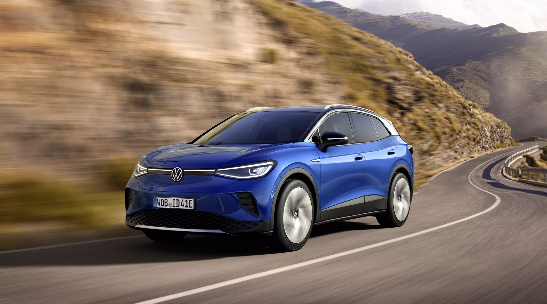 Dynamisk vy av en blå VW ID4 1:a generationen från 2020, som visar fram- och sidoprofiler med LED-strålkastare, aerodynamisk design, och legeringshjul på en kurvig väg, vilket antyder avancerad teknologi samt elektriska eller hybrida förmågor