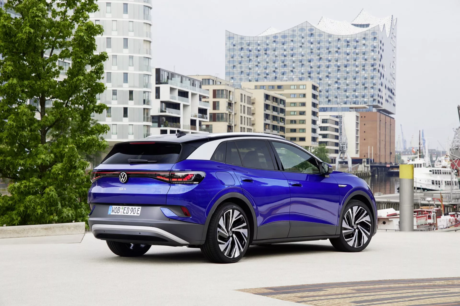 VW ID.4 1:a generationen 2020 fotograferad från ett bakifrån tre fjärdedels perspektiv som visar dess bakre designelement inklusive LED-bakljus, bakre stötfångare med integrerade parkeringssensorer och emblem, tillsammans med dess aerodynamiska sidoprofil med stora flerspakiga legeringshjul, dörrhandtag i karossfärg och tonat glastak eller panoramatak inställt mot en urban bakgrund med modern arkitektur.