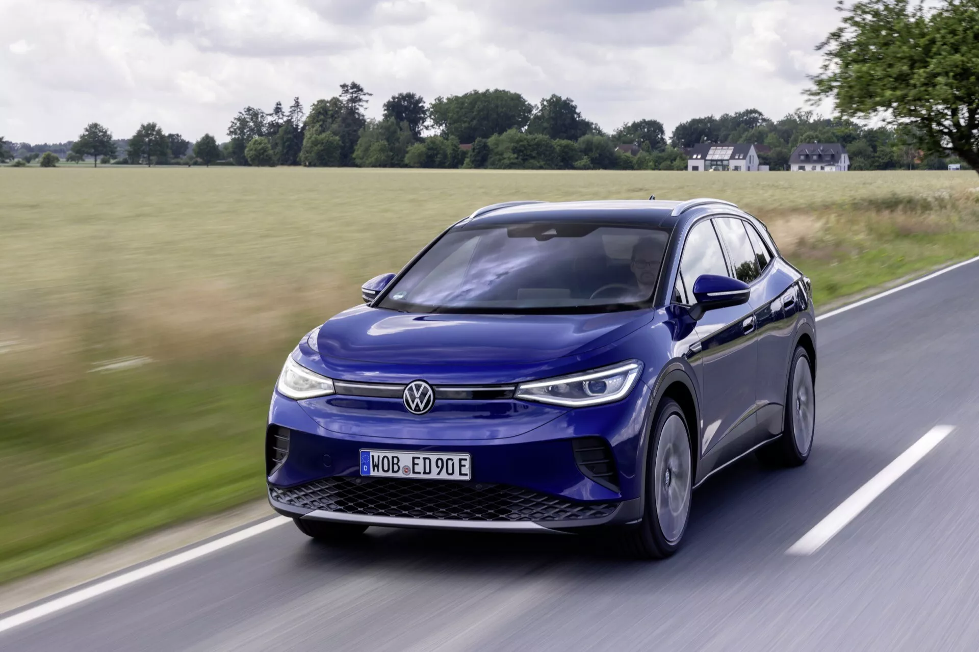 Blå Volkswagen ID.4 elektrisk SUV i rörelse, sedd från ett lågt framåt trekvartsperspektiv som framhäver dess eleganta LED-strålkastare, stängda grill, legeringshjul och dynamiska designlinjer mot en lantlig bakgrund.