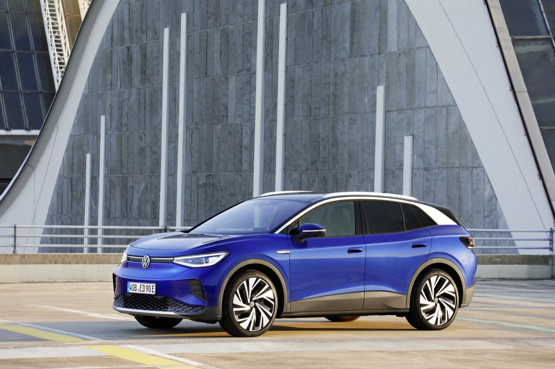 Blå Volkswagen ID.4 2020:s sida profil med aerodynamisk design, släta horisontella strålkastare, flerspakiga legeringshjul och LED-indikatorer mot en modern urban bakgrund som visar geometriska strukturer och vertikala linjer