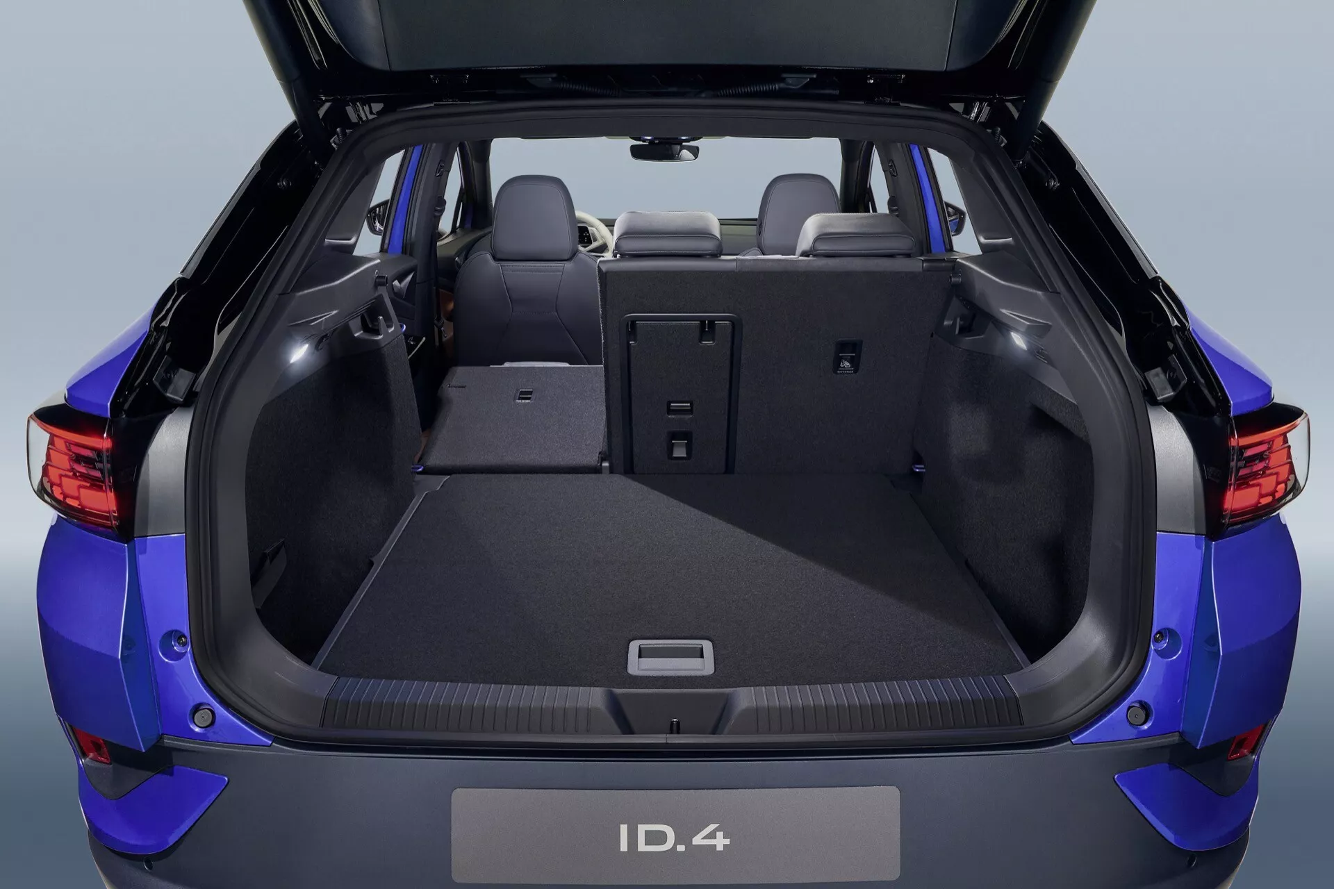 Baksida av en Volkswagen ID.4 2020 som visar öppet bagageutrymme med detaljerat lastutrymme och moderna funktioner som ett 12V-uttag, platt lastgolv täckt med matta, 60/40 delbara baksäten och integrerad spoiler med tredje bromsljus