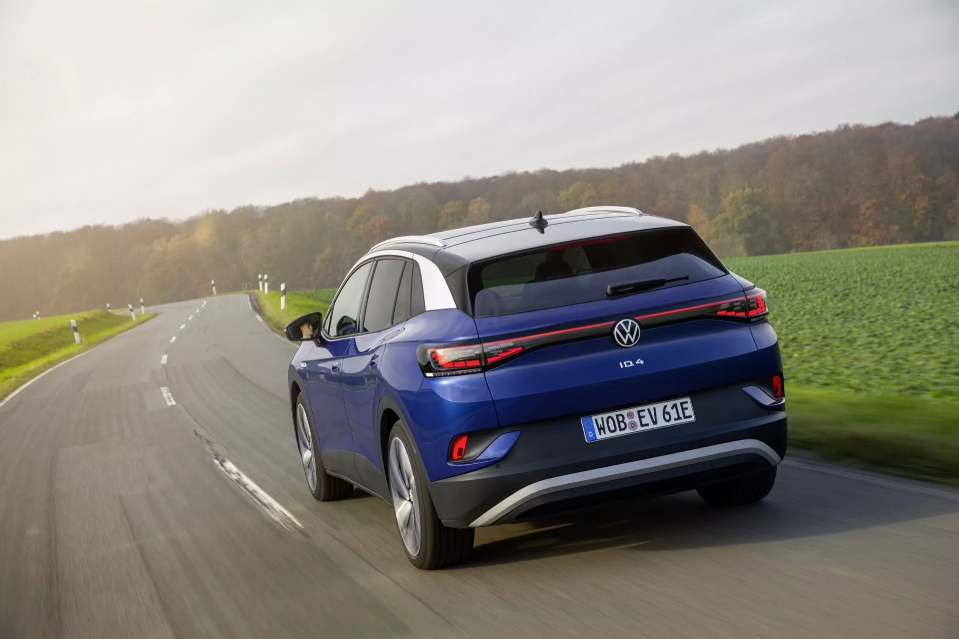 Dynamisk bakre tre fjärdedels vy av en blå Volkswagen ID.4 elektrisk SUV i rörelse på en asfalterad väg, med LED-baklyktor, takspoiler, lysande yttre och synlig registreringsskylt med 'E'-beteckning, vilket anger status som elektriskt fordon.