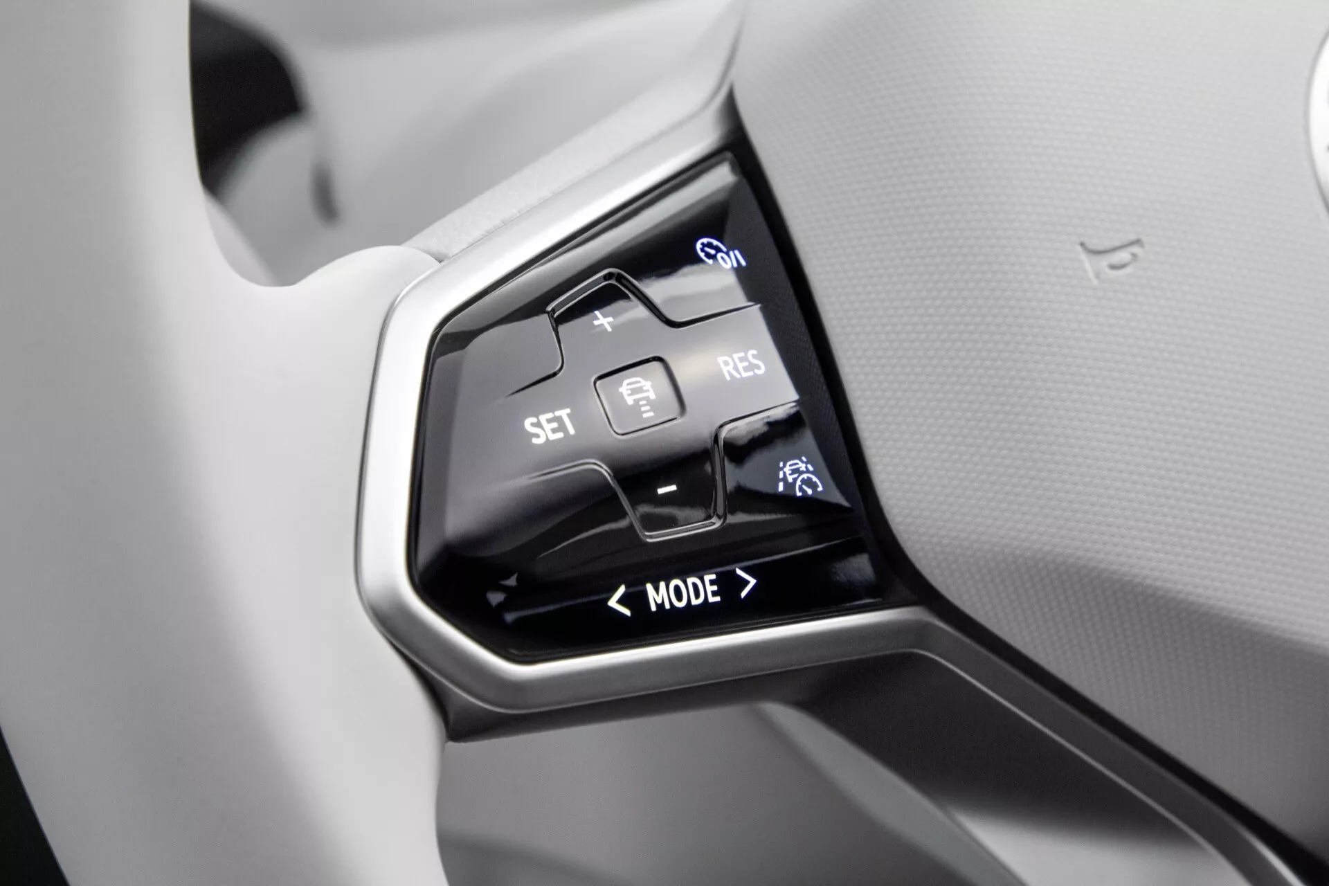 Närbild av styrkontroller på ratten i VW ID4 första generationen 2020 med fokus på adaptiv farthållare, körningslägen och röstkommandofunktioner på en blank svart panel mot en ljusgrå ratt.
