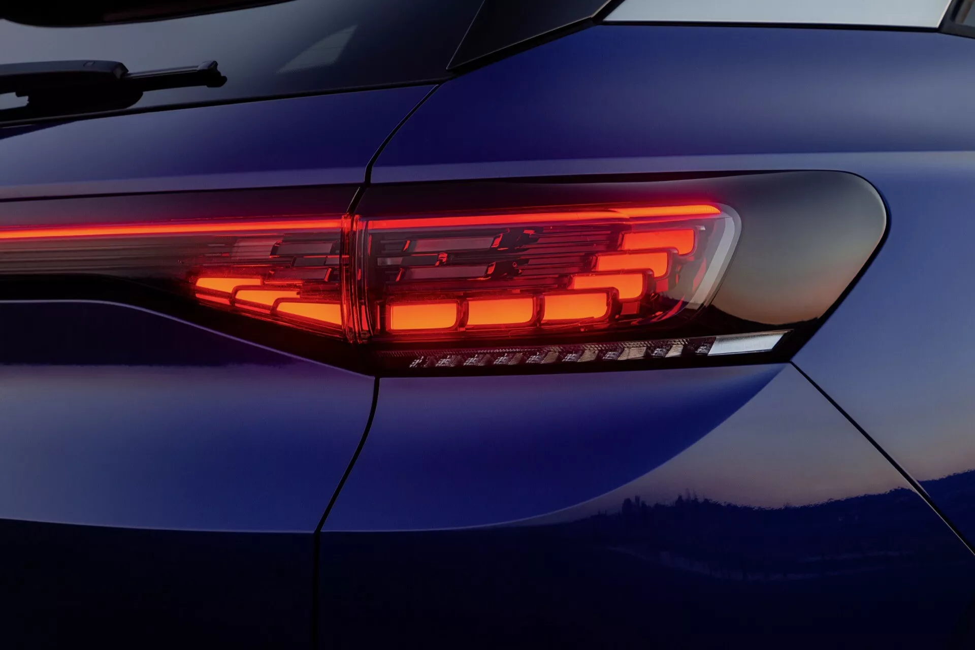 Närbild av den moderna LED-baklyktedesignen på 2020 års VW ID.4 1:a generation som visar dess snygga horisontella remsa och komplexa upplysta segment mot en reflekterande djupblå kaross