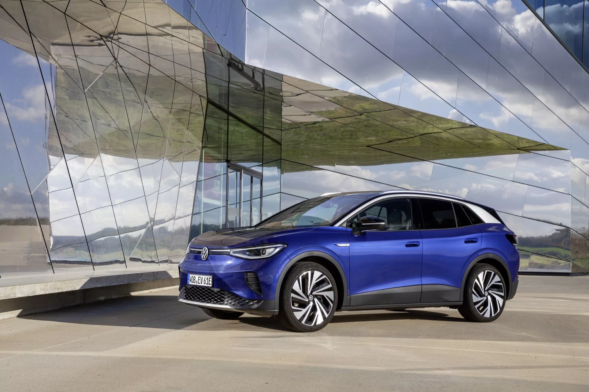 VW ID.4 1:a generationen 2020 i levande blått, med modern SUV-design från en trekvarts framsida på slät betong, mot en vinklad arkitektonisk bakgrund med glas och metall, visar LED-strålkastare, unika legeringshjul och en slank sidoprofil.