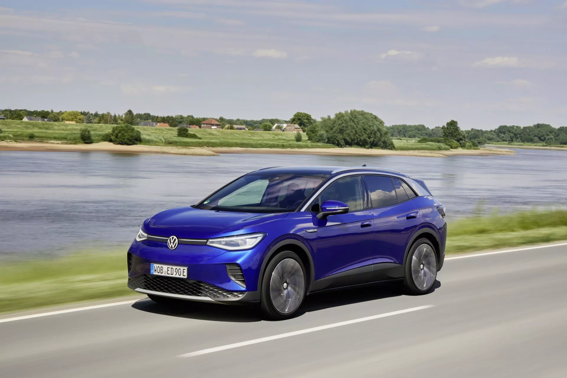 Dynamisk trekvarts framsida av den elektriska bilen VW ID.4 2020 i livligt blått, som visar dess slanka design och rörelse mot en naturlig motorvägsbakgrund med en lugn vattenkropp och grönska