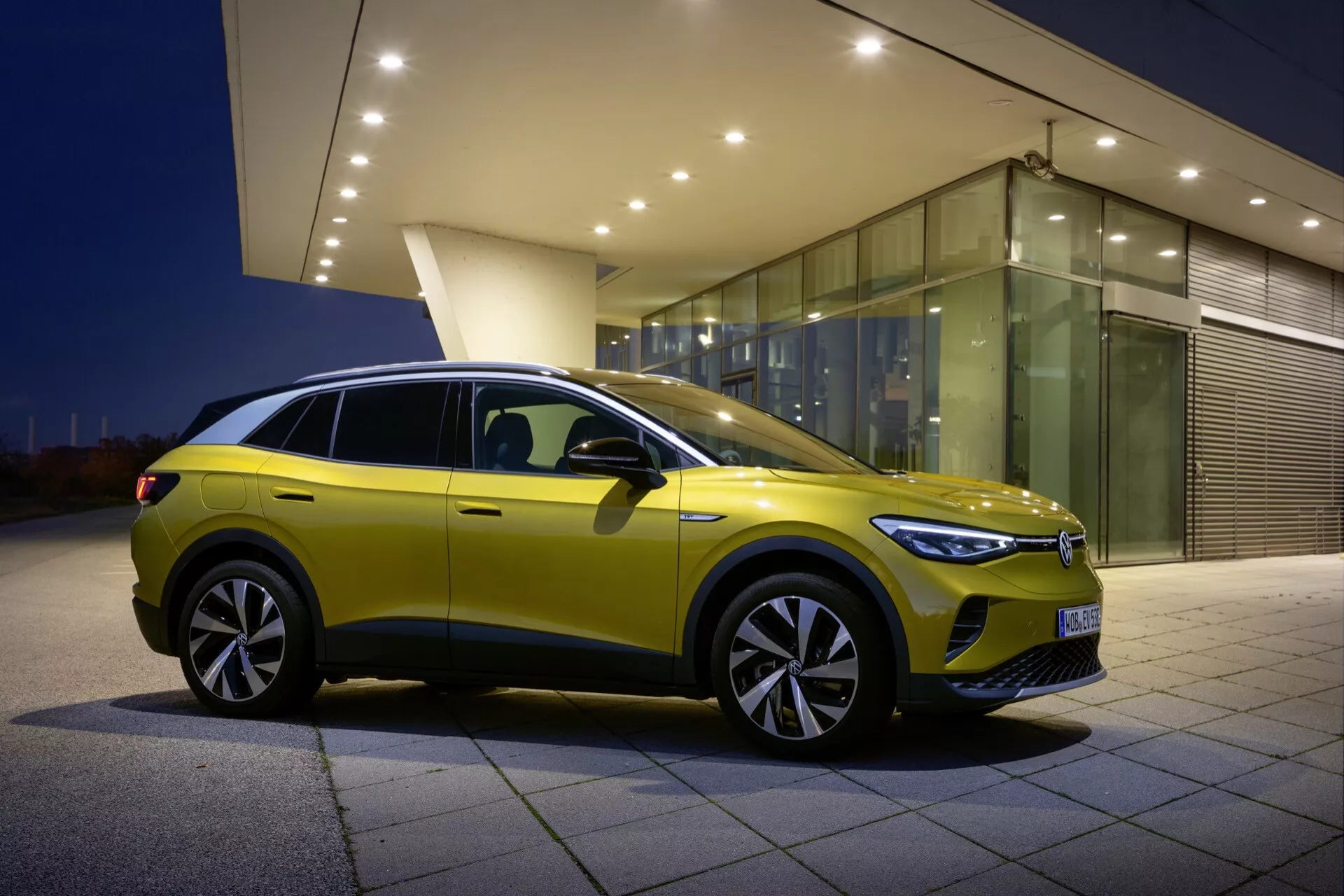 VW ID.4 första generationen 2020 i livligt gult med vy från framsidan och sidan, framhäver dess LED-strålkastare, kraftiga kaross och stora legeringshjul mot en urban skymningsbakgrund