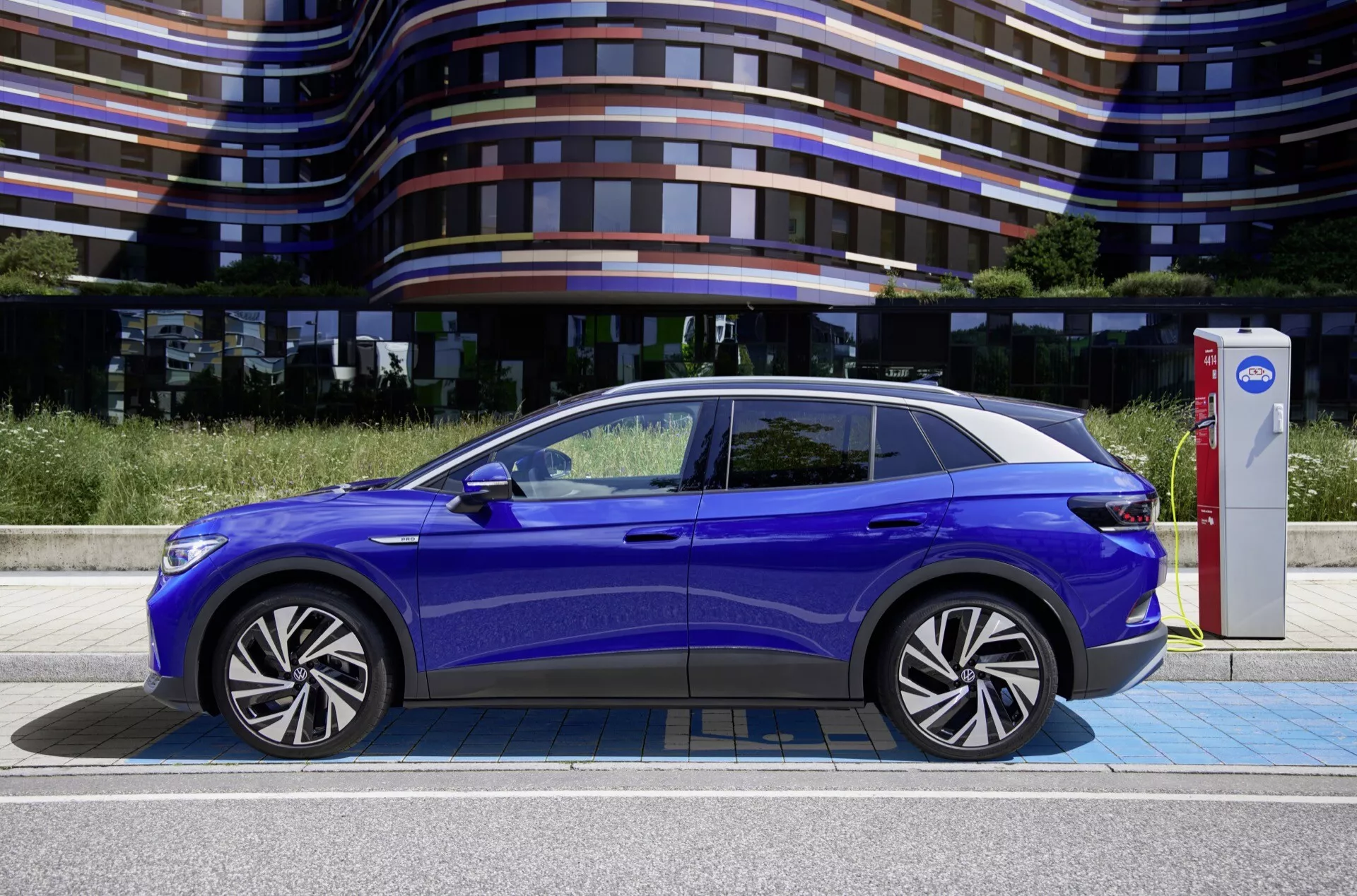 Profilvy av en blå 2020 VW ID.4 elbil, som belyser dess sidodesignelement, från de eleganta LED-strålkastarna och stängda grillen framtill, via de infällda dörrhandtagen och konturerade karosslinjerna, till det panoramiska glastaket och legeringshjulen, med bilen ansluten till en laddningsstation.