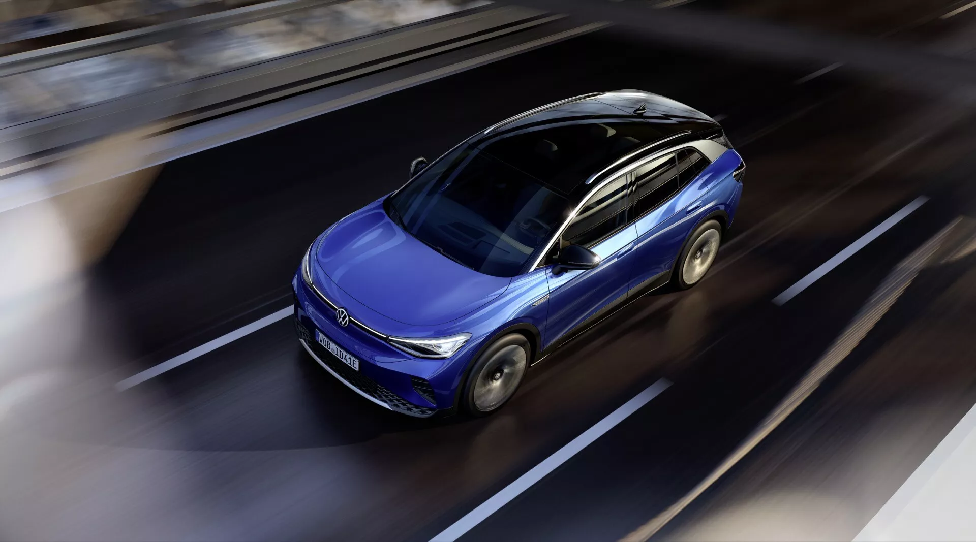 Dynamisk fågelperspektiv av en modern blå VW ID.4 2020 i rörelse, som framhäver dess slanka design, soltak, konturerade huva och aerodynamiska kaross mot en suddig vägbakgrund, antydande avancerade funktioner och miljövänlig teknik i dagsljus.