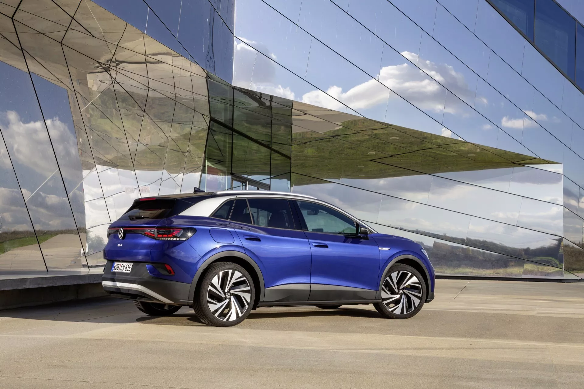 VW ID.4 1:a Generationen 2020 i Metallisk Blå - Bakifrån och Tre Fjärdedels Vy som visar Aerodynamisk Design, Sportigt Ställning, Stora Legeringshjul, Moderna LED Bakljus, och Premium Finish mot en Arkitektonisk Bakgrund
